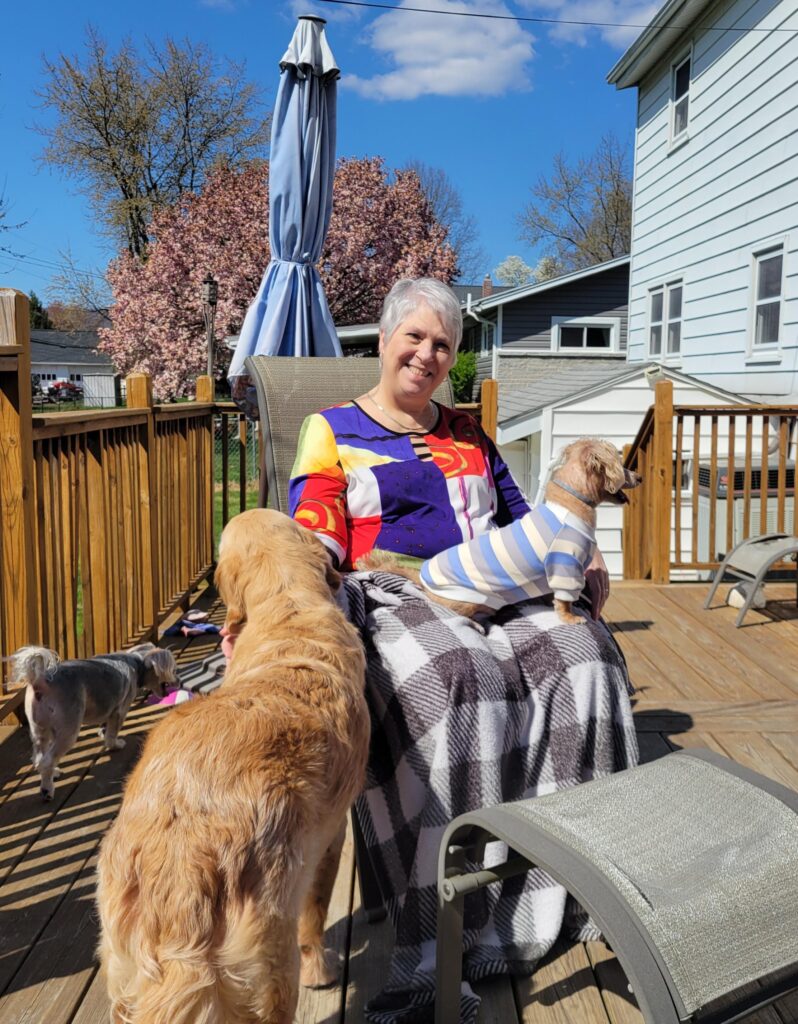 Lynn Brakefield sitting on a sunny deck with three dogs, including one in a striped sweater, surrounded by spring blossoms and blue skies.