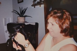 Woman with short auburn hair smiling and holding a small black kitten indoors, with a wooden table, potted plant, and decorative eagle figurines in the background.